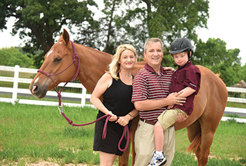 a woman, a man, and a little boy held by the man while wearing a riding helmet pose in front of a horse in front of a fence